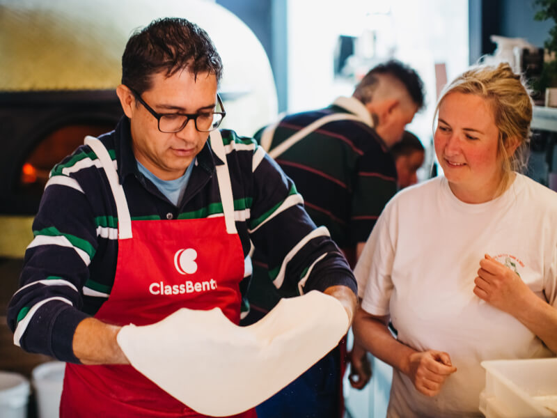 Woman showing man wearing apron how to make pizza. 